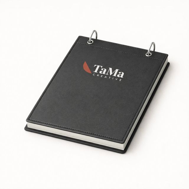 Bloco de mesa preto com capa pespontada e duas argolas prateadas no topo, com a marca TaMa em branco, folha em vermelho, na capa.