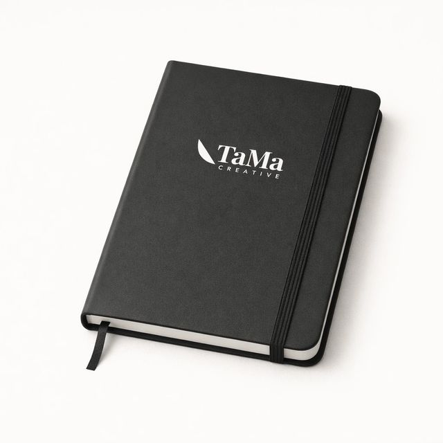 Caderno A5 preto com elástico e marcador de página, com a marca TaMa em branco na capa.