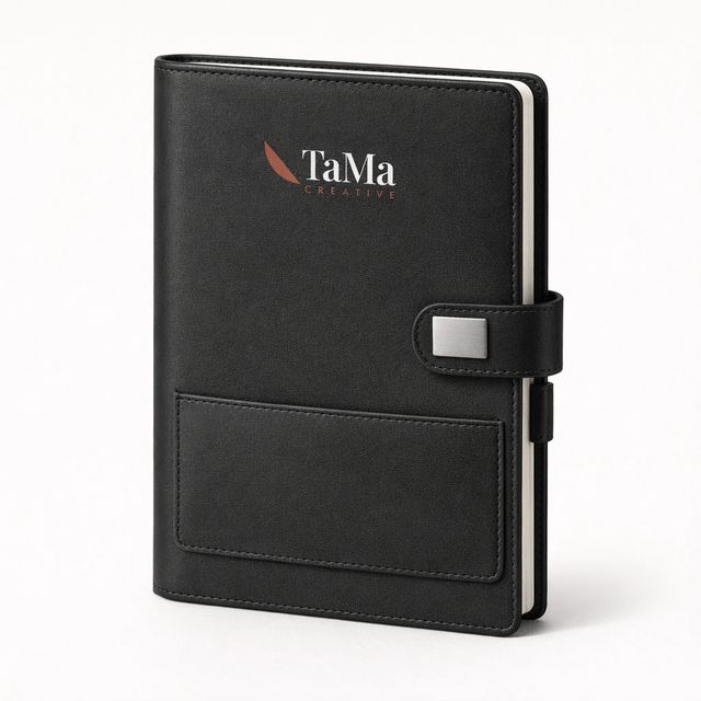 Caderno preto com aba de fechamento com plaquinha prata, bolso porta-cartão e alça para caneta, com a marca TaMa em branco e vermelho na capa.