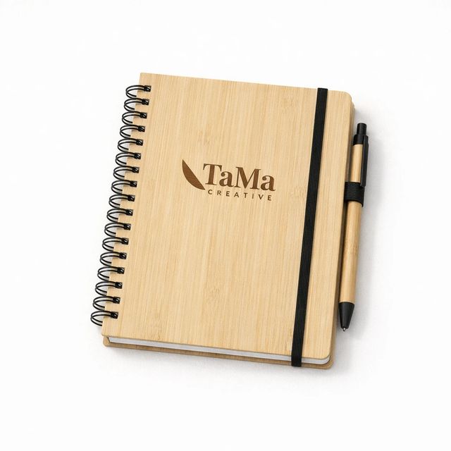 Caderno com capa em tom natural, espiral preto, elástico preto e caneta em tom natural, com a marca TaMa gravada em marrom na capa.