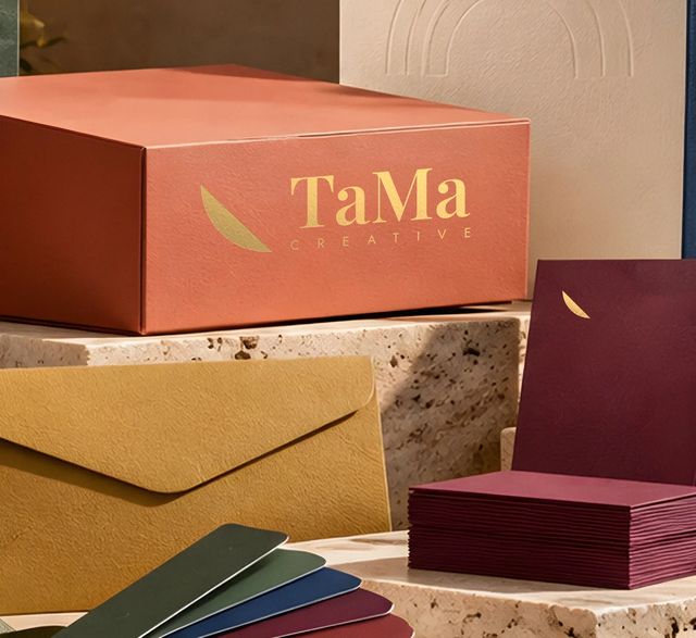 Caixa rígida terracota com logo TaMa Creative em hot stamping dourado na face frontal e cartão bordô com pétala dourada. Mockup ilustrativo.