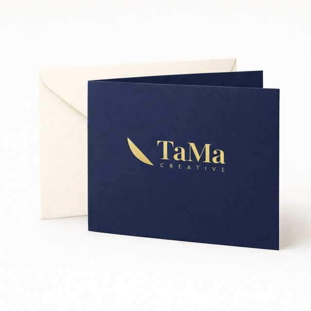 Cartão dobrado azul-marinho ao lado de um envelope creme, com a marca TaMa em dourado.