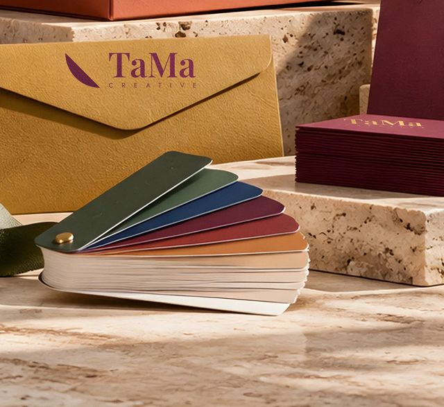 Envelope mostarda com logo TaMa Creative em hot stamping ameixa, pilha de cartões bordô com TaMa Creative dourado e leque de cores. Mockup ilustrativo.
