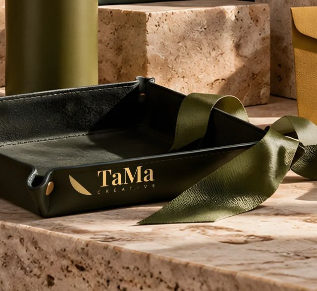 Bandeja de couro verde-escuro com a assinatura TaMa Creative em hot stamping dourado na lateral, envolta por uma fita verde-oliva sobre travertino. Mockup ilustrativo.