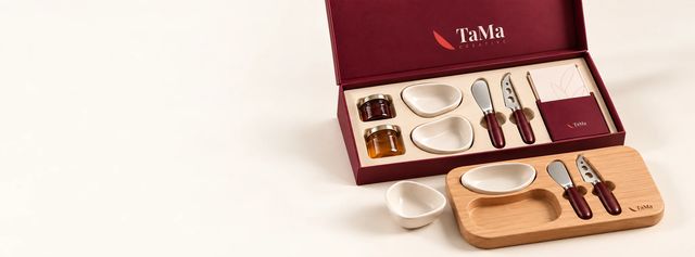 Caixa bordô aberta com tábua de madeira, potinhos de cerâmica, geleias, facas de queijo e cartão com a marca TaMa.