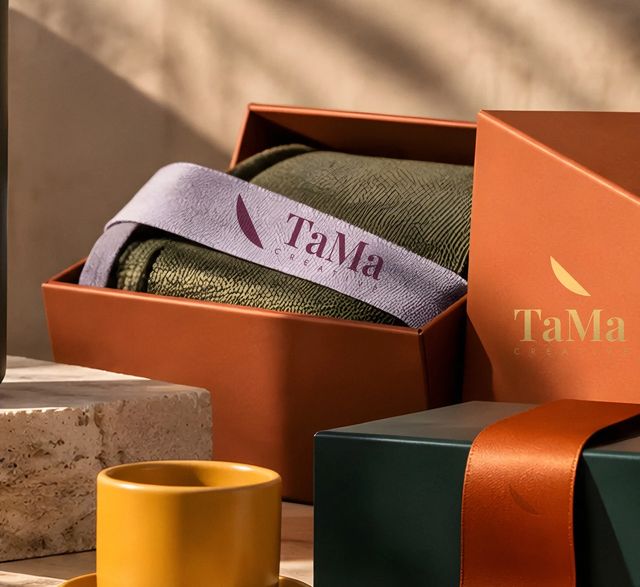 Manta oliva em caixa terracota com faixa lilás estampada com a assinatura TaMa Creative em ameixa. Mockup ilustrativo.