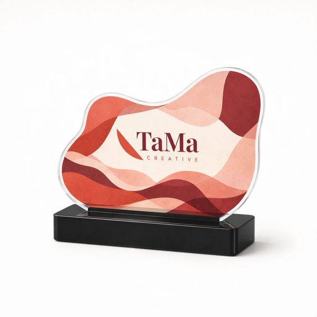 Troféu de placa transparente recortada no contorno de uma ilustração abstrata em tons de vermelho e rosa, em base preta, com a marca TaMa impressa na placa.